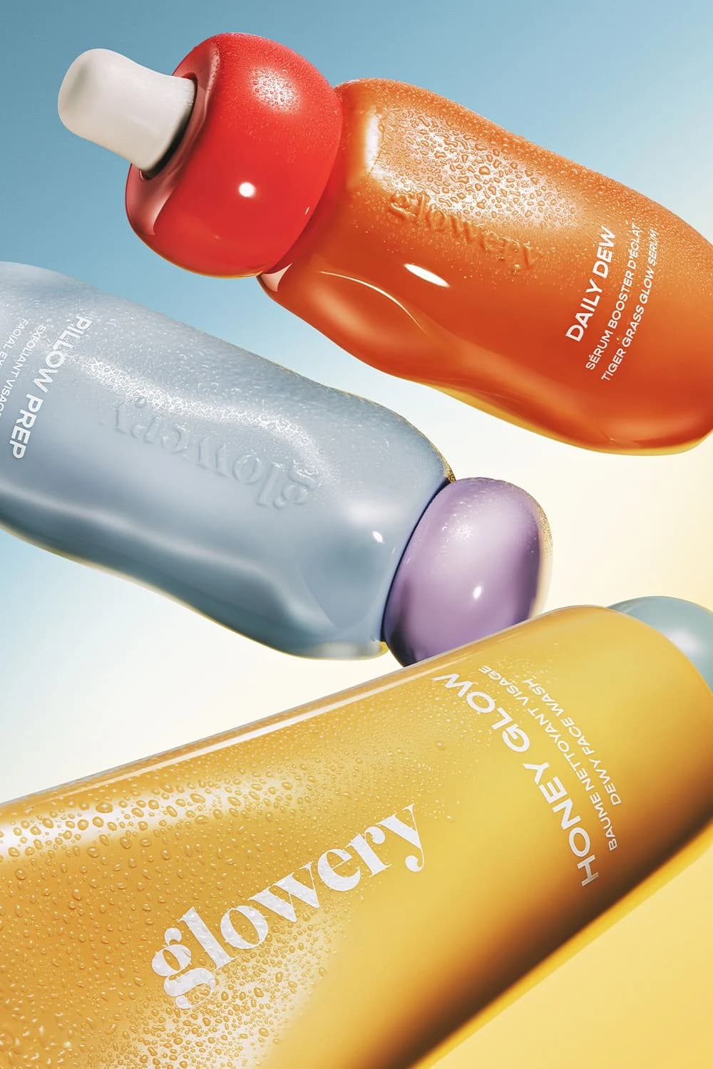 Three glossy skincare bottles in orange, pale blue and yellow tumbling diagonally across a gradient background from sky blue to cream, covered in fine condensation with soft coloured caps
