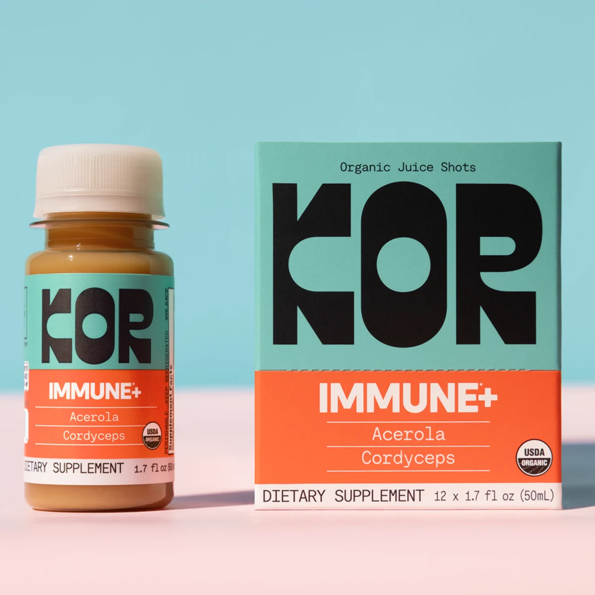 A KOR Immune Plus juice shot bottle standing beside its matching carton on a pink surface against a pale blue background, both showing the KOR wordmark, the ingredient lines and a USDA Organic seal